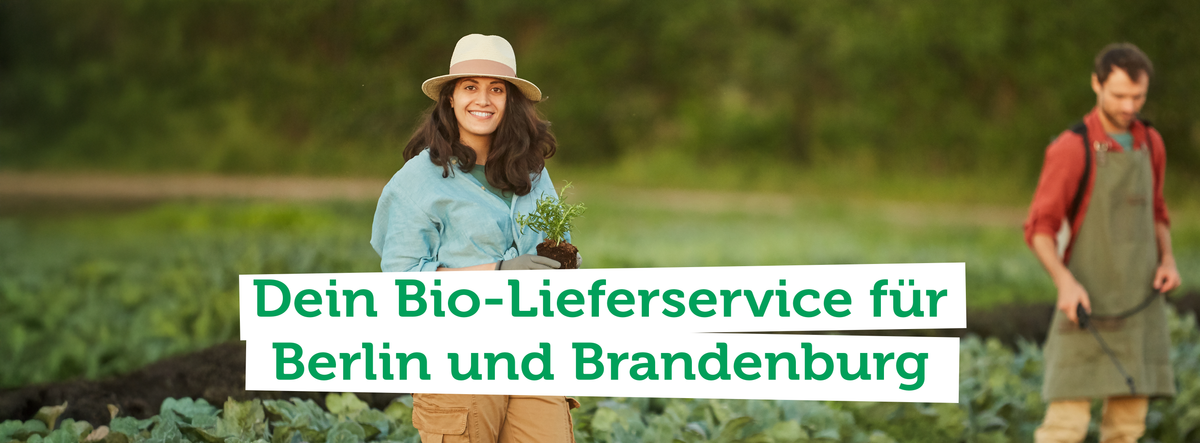 Eine junge Frau mit Hut und Arbeitskleidung und ein junger Mann in Arbeitskleidung mit Schürze stehen in einem Gemüsefeld. Die Frau schaut direkt in die Kamera und hält eine Jungpflanze in den Händen.