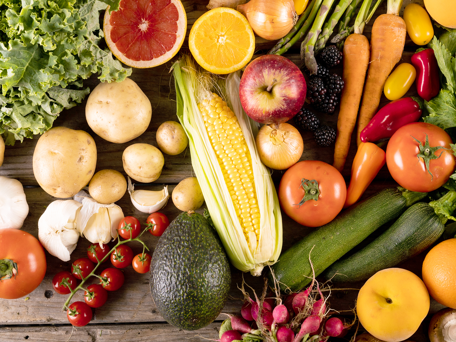 Vogelperspektive auf eine Holzfläche auf der bunt gemischtes Obst und Gemüse liegt, wie Mais, Zucchini, Möhren, Orangen und Radieschen