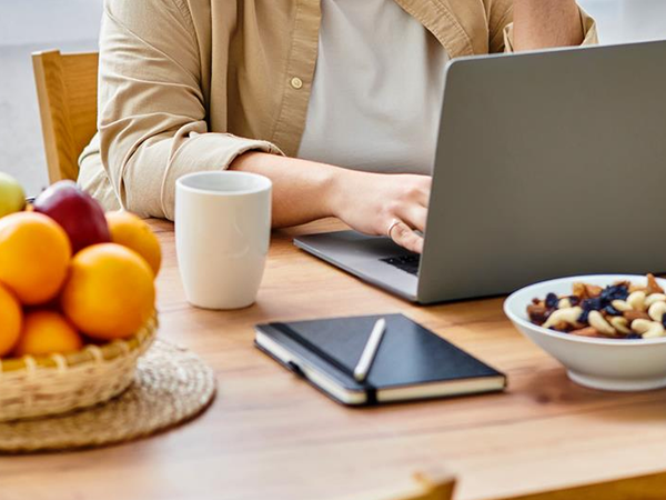 KI generiert: Eine Person arbeitet an einem Laptop am Tisch mit Obst, einer Tasse, Notizbuch und einer Schale Nüsse.