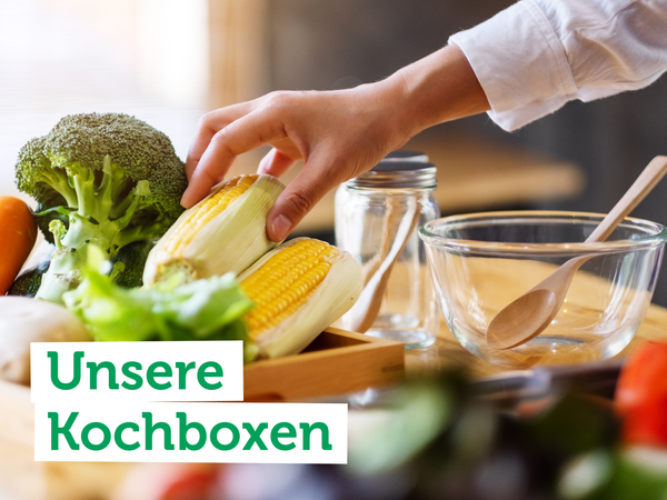 Hand greift nach Mais, umgeben von Gemüse, Schalen und Kochutensilien. Text auf dem Bild: "Unsere Kochboxen".