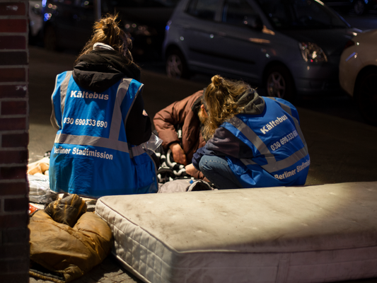 KI generiert: Zwei Personen im "Kältebus"-Westen helfen einer obdachlosen Person auf der Straße.