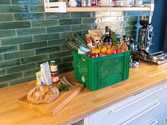 KI generiert: Grüne Kiste mit Lebensmitteln und Brot auf einem Holztresen in einer Küche.