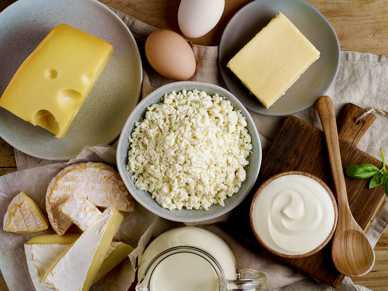Vogelperspektive auf eine Holzfläche auf der verschiedene Molkereiprodukte wie Käse am Stück, Eier und eine Schale Joghurt angeordnet sind.