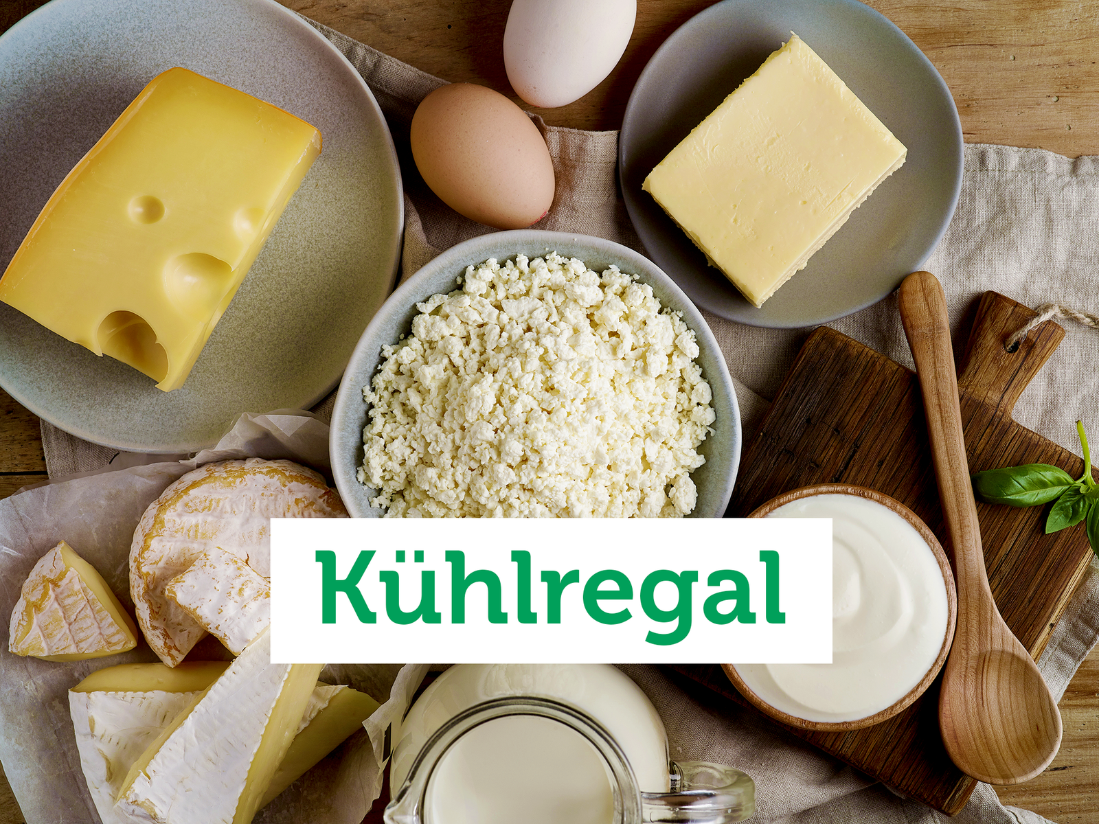 Vogelperspektive auf eine Holzfläche auf der verschiedene Molkereiprodukte wie Käse am Stück, Eier und eine Schale Joghurt angeordnet sind.
