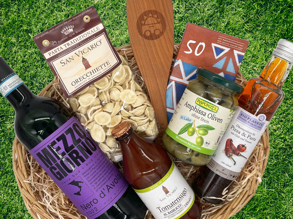 KI generiert: Ein Geschenkkorb mit Wein, Pasta, Oliven, Tomatensugo, Schokolade und Öl auf grünem Hintergrund.