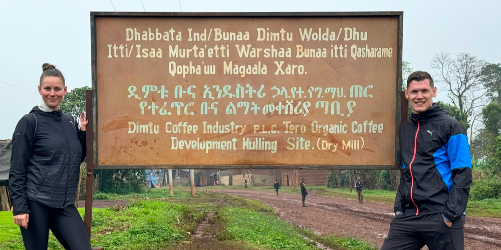 KI generiert: Zwei Personen stehen neben einem Schild. Text auf dem Schild: "Dimtu Coffee Industry PLC, Tero Organic Coffee Development Hulling Site (Dry Mill)."