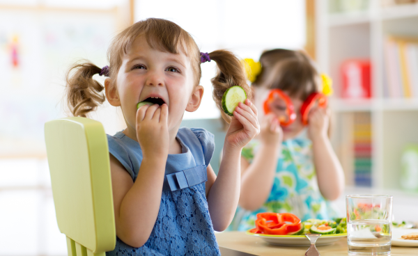 KI generiert: Zwei Kinder essen Gemüse an einem Tisch; eines hält eine Gurkenscheibe, das andere spielt mit Paprika.
